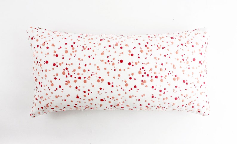Op de afbeelding: Een wit rechthoekig kussen met een roze en rood splatterpatroon.