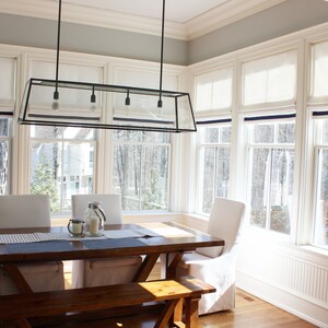 May include: A dining room with a wooden table and chairs, a bench, and a large glass and metal chandelier. The room has large windows with white roman shades and a view of trees and a backyard.