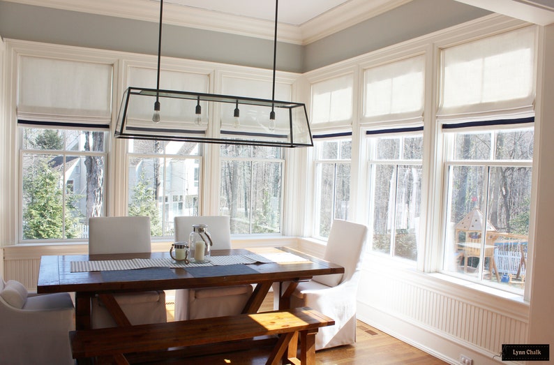 May include: A dining room with a wooden table and chairs, a bench, and a large glass and metal chandelier. The room has large windows with white roman shades and a view of trees and a backyard.
