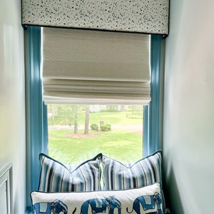 May include: A window with a blue frame and a white valance with a blue and white polka dot pattern. The window has a white roman blind and a view of a green lawn. There are three blue and white striped cushions and a white cushion with blue elephants on a window seat.