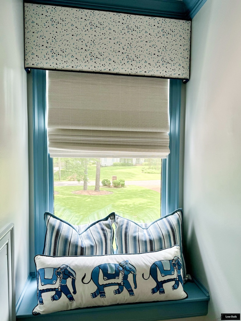 May include: A window with a blue frame and a white valance with a blue and white polka dot pattern. The window has a white roman blind and a view of a green lawn. There are three blue and white striped cushions and a white cushion with blue elephants on a window seat.