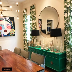 Może przedstawiać: Zielona komoda z dużym okrągłym lustrem nad nią. Komoda ma drewniany blat i jest ozdobiona białymi choinkami i kulami śnieżnymi. Pok&oacute;j ma zielone zasłony z białym wzorem liści i duży abstrakcyjny obraz na ścianie.