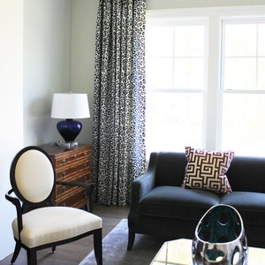 May include: A living room interior featuring a black sofa, a white chair, a glass coffee table, and a lamp with a blue base. The sofa has a red and gold geometric patterned cushion. The room has a grey rug and white walls with a white and black patterned curtain.
