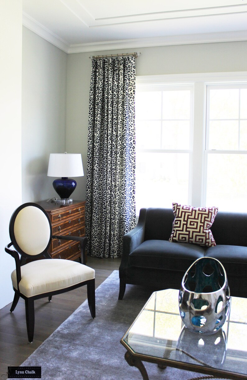 May include: A living room interior featuring a black sofa, a white chair, a glass coffee table, and a lamp with a blue base. The sofa has a red and gold geometric patterned cushion. The room has a grey rug and white walls with a white and black patterned curtain.