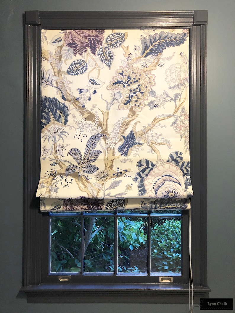 May include: A white roman shade with a floral pattern in shades of blue, purple, and gold. The shade is pulled up to reveal a window with a view of green foliage.