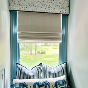 Puede incluir: Un asiento de ventana con almohadas a rayas azules y blancas y una almohada larga blanca con elefantes azules. La ventana tiene una cenefa blanca con lunares azules y una cortina romana blanca.