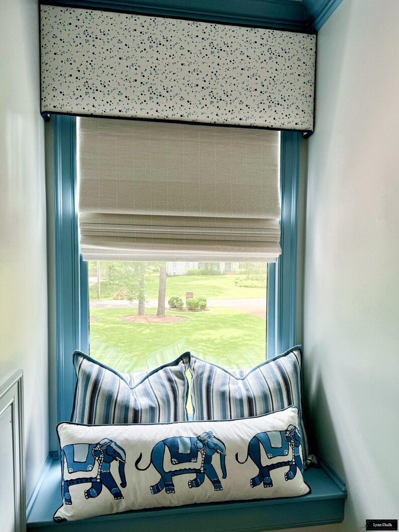 Puede incluir: Un asiento de ventana con almohadas a rayas azules y blancas y una almohada larga blanca con elefantes azules. La ventana tiene una cenefa blanca con lunares azules y una cortina romana blanca.