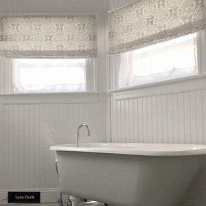 Op de afbeelding: Een witte badkamer met een bad op pootjes en witte lambrisering. De ramen hebben witte rolgordijnen met een grijs en wit bladmotief.