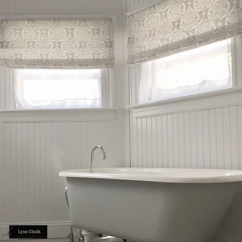 Op de afbeelding: Een witte badkamer met een bad op pootjes en witte lambrisering. De ramen hebben witte rolgordijnen met een grijs en wit bladmotief.