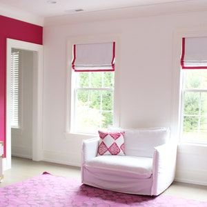 May include: A white upholstered armchair with a pink patterned pillow sits in a room with two windows with white roman shades trimmed in pink. The room has a pink wall and a pink rug.