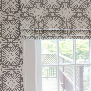 May include: A white window with a gray and white patterned roman shade. The shade is pulled up and the window is open, showing a view of a wooden deck and green trees.