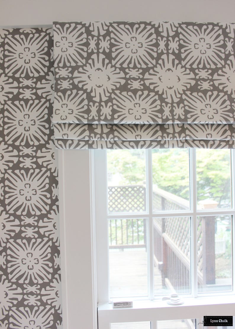 May include: A white window with a gray and white patterned roman shade. The shade is pulled up and the window is open, showing a view of a wooden deck and green trees.