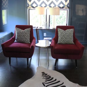 May include: Two red velvet armchairs with black and white patterned cushions sit in front of a window with a black and white patterned valance. A zebra print rug is on the floor in front of the chairs.