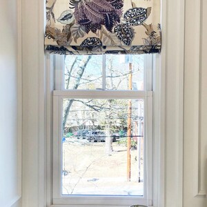 May include: A white window with a floral patterned roman shade. The shade has a cream background with large purple and blue flowers. The window is framed by white trim and is set against a white wall. A white lamp with a white shade sits on a white surface below the window.