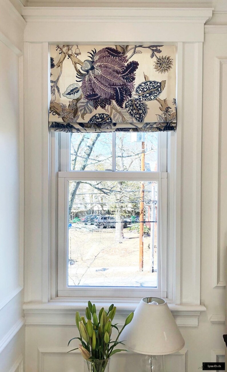 May include: A white window with a floral patterned roman shade. The shade has a cream background with large purple and blue flowers. The window is framed by white trim and is set against a white wall. A white lamp with a white shade sits on a white surface below the window.
