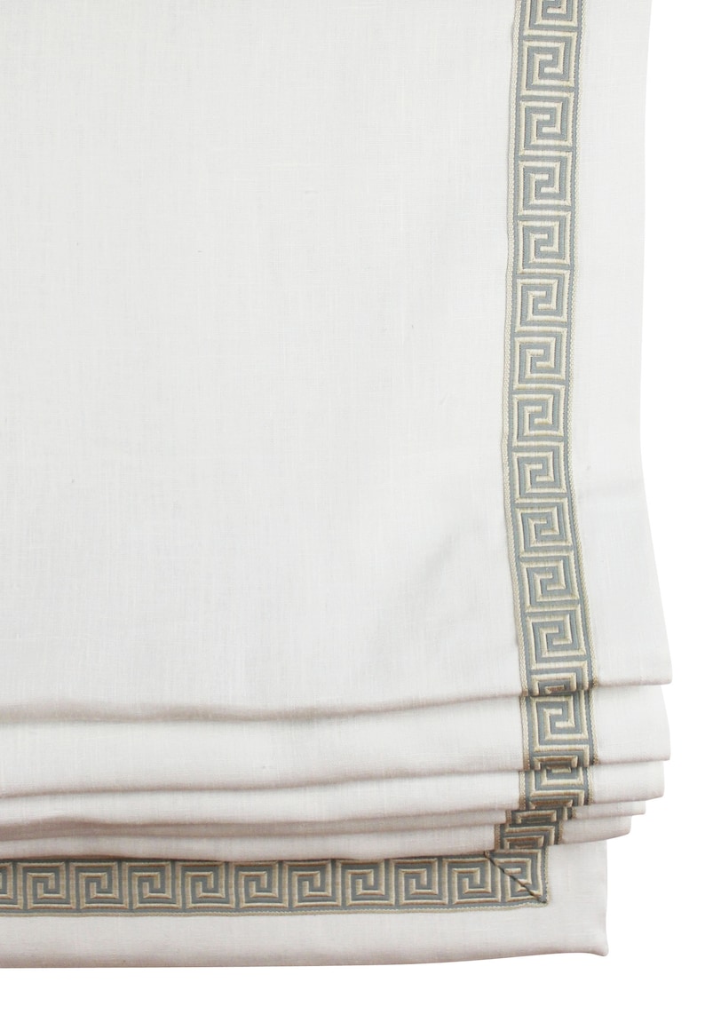 May include: White roman shade with a gray and cream greek key trim. The shade is folded up, showing the layers of fabric.