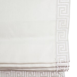 May include: White roman shade with a white greek key border. The shade is rolled up and the bottom edge is visible.