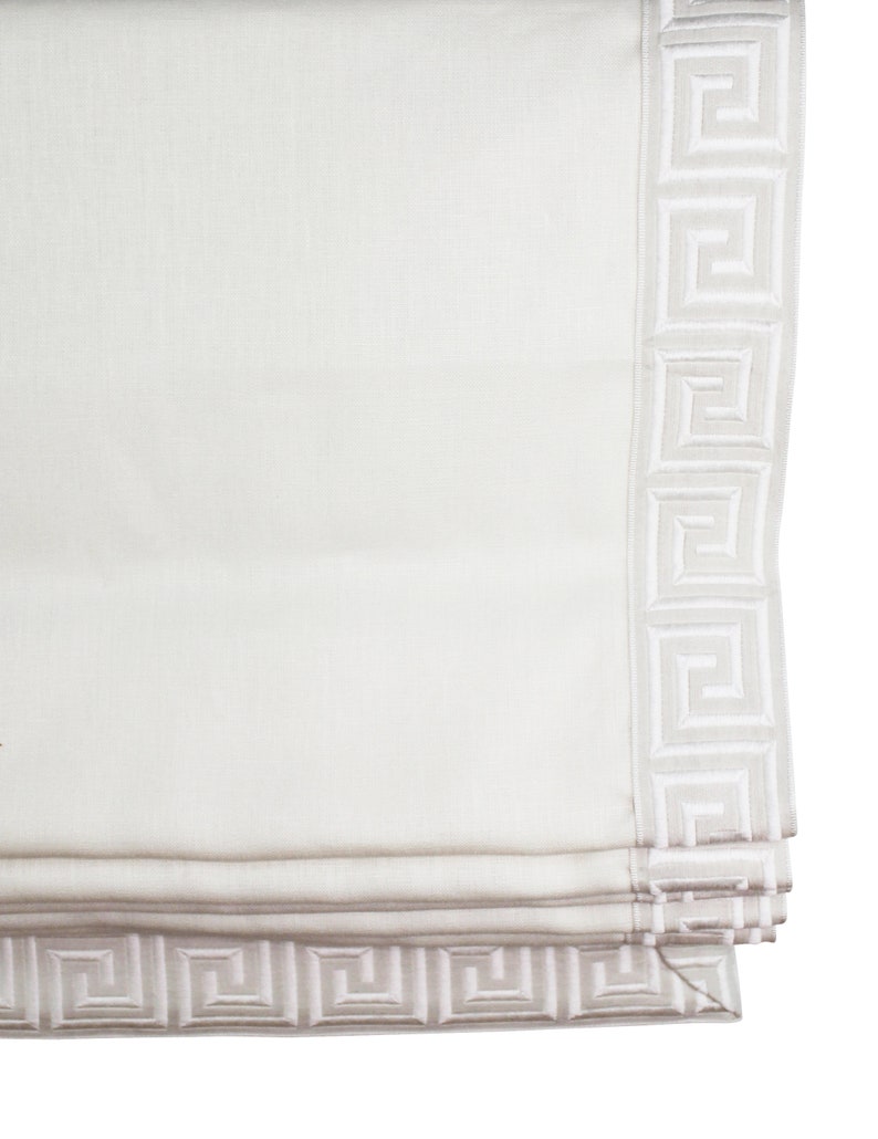 May include: White roman shade with a white greek key border. The shade is rolled up and the bottom edge is visible.
