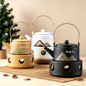 May include: Three ceramic teapots in beige, white, and black, each with a matching warmer. The teapots have mountain landscape designs and text that reads "Nana's Pot", "Tea & Love", and "Family Tea". Each teapot has a metal handle.