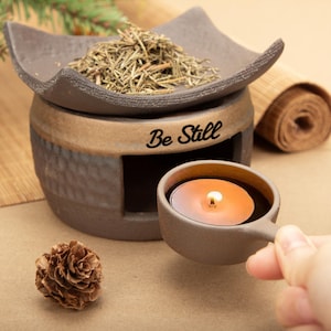 May include: A brown ceramic aromatherapy diffuser with a candle and the text "Be Still". The diffuser has a tray for herbs and a small cup with a lit candle. A pine cone is in the foreground.