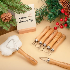 Peut inclure: Un ensemble d'outils de poterie avec des manches en bois et des lames en métal. Les outils sont disposés sur une surface claire, avec une carte indiquant "Pottery Lover's Gift". Les outils sont étiquetés "Clay & soul" et "Create with love".