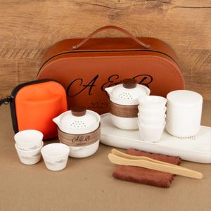 May include: A complete tea set with a brown leather carrying case, white ceramic teacups, a teapot, and a tea tray. The set includes a small orange zippered pouch, wooden tongs, and a brown cloth. The case is embossed with the letters "A & B" and the year "2025".