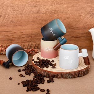 May include: A collection of ceramic mugs with unique designs. One mug is black and blue with the name "Mabel", another is white with "M & K", and a third is pink with the word "Love". Each mug has a wooden handle.