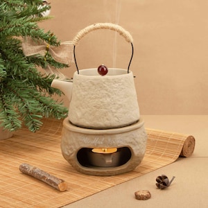 Peut inclure: Un chauffe-huile en céramique beige en forme de théière, avec une poignée en corde et un bouton rouge. La base comporte un compartiment pour une bougie chauffe-plat, avec l'inscription "Mindful Home". De la vapeur s'élève du haut. Un petit sapin de Noël est en arrière-plan.