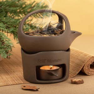May include: A brown ceramic aromatherapy diffuser with a candle and the text "Still Moment". The diffuser has a teapot-like design with a handle and spout. Dried herbs sit in the top, with steam rising. A lit candle sits below.