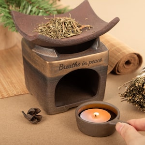 May include: A brown ceramic essential oil diffuser with a square base and a curved top. The words "Breathe in peace" are inscribed on the front. A lit tea light candle sits in a small cup. Dried herbs are on the top.