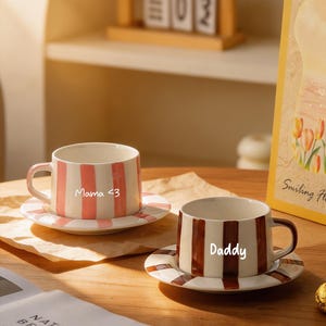 Puede incluir: Dos tazas de café de cerámica con platillos a juego. Una taza tiene rayas rosas y blancas y el texto "Mama <3". La otra taza tiene rayas marrones y blancas y el texto "Daddy". Ambas tazas están sobre una mesa de madera.