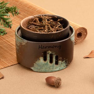 May include: A ceramic tea warmer with a brown and teal glaze. The warmer has three rectangular cutouts and the word "Harmony" printed on the front. A small bowl filled with dried tea leaves sits inside the warmer.