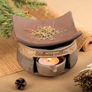May include: A brown ceramic essential oil diffuser with a square dish on top holding dried herbs. The base has a candle holder with a lit tea light. The text "Stillness within" is inscribed on the front. A pine cone and greenery are in the background.