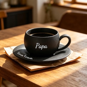 May include: A black ceramic coffee cup and saucer set. The cup is filled with dark coffee and has the word "Papa" printed on the side. The saucer has the words "Since Forever" printed on it. The set is on a wooden table.