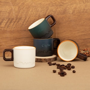 Op de afbeelding: Vier keramische koffiemokken met bruine handvatten en randen. De mokken zijn in tinten wit, blauwgroen en blauw, met het woord "Coffee" en andere tekst. Koffiebonen liggen verspreid bij de mokken.