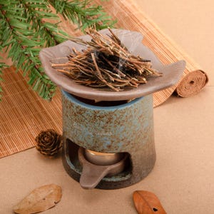 May include: A ceramic essential oil diffuser with a brown leaf-shaped dish holding dried herbs that are smoking. The diffuser is blue and brown with a small opening for a tea light. A pine cone and leaves are nearby.