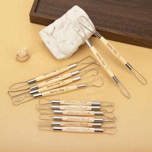 May include: A collection of pottery tools with wooden handles and metal loops. The tools are labeled "Sienna Clay" and "Artisan's Touch". A piece of clay and a small clay sculpture are also present.