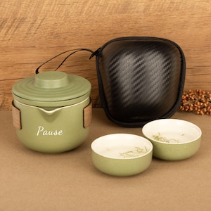 May include: A green ceramic tea set with a matching travel case. The set includes a lidded teapot with the word "Pause" and two small bowls. The travel case is black with a textured pattern.