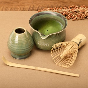 Könnte beinhalten: Ein Matcha-Teeset mit einer grünen Keramikschale mit Matcha-Tee, einem Schneebesen, einem Löffel und einer Keramik-Teetasse. Auf der Schale steht das Wort "Calm". Das Set befindet sich auf einer braunen Oberfläche.