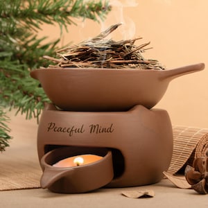 Puede incluir: Un difusor de aromaterapia de cerámica marrón con asa, que contiene hierbas secas que emiten humo. Debajo hay una vela encendida. Las palabras "Peaceful Mind" están inscritas en la parte delantera. El difusor está diseñado para aromaterapia.