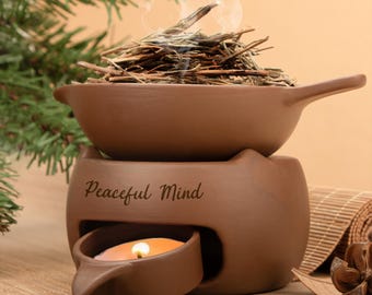 Puede incluir: Un difusor de aromaterapia de cerámica marrón con asa, que contiene hierbas secas que emiten humo. Debajo hay una vela encendida. Las palabras "Peaceful Mind" están inscritas en la parte delantera. El difusor está diseñado para aromaterapia.
