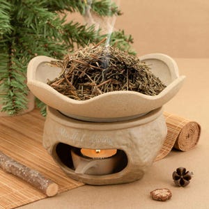 May include: A beige ceramic essential oil diffuser with a floral-shaped top, filled with dried herbs. A lit candle sits inside the base, with the words "Mindful Living" etched on the front. A small sprig of evergreen and a rolled bamboo mat are in the background.