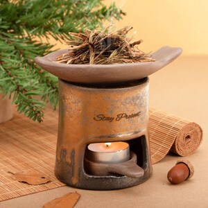 Puede incluir: Un difusor de aceites esenciales de cerámica en tonos marrón y dorado. El difusor tiene una bandeja para hierbas, un portavelas y el texto "Stay Present". Hay una vela de té encendida dentro.