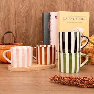 Può includere: Una collezione di tazze in ceramica a righe in vari colori, tra cui rosa, rosso, nero e verde. Le tazze sono disposte su una superficie di legno con libri e oggetti decorativi sullo sfondo. Una tazza ha il testo "But First Coffee".