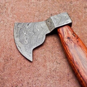 May include: A small hatchet with a patterned steel blade and a wooden handle. The blade has a unique swirling design, and the handle is made of reddish-brown wood. The hatchet is set against a textured, reddish-brown background.