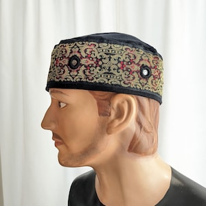 May include: Black velvet kufi with a gold and red embroidered band. The band has a repeating pattern and features two black buttons.