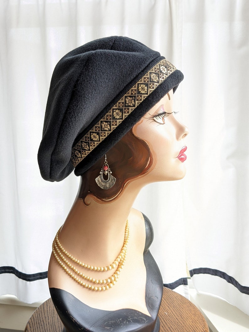 Puede incluir: Un gorro de lana negro con un borde de patr&oacute;n dorado y negro. El gorro se lleva en un maniqu&iacute; con un collar de perlas y pendientes.