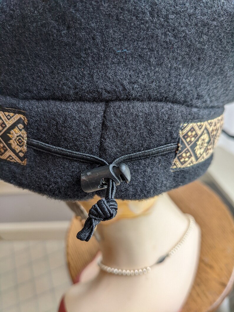 Puede incluir: Sombrero de forro polar negro con una banda estampada en oro y negro y una correa ajustable negra con una hebilla de pl&aacute;stico.