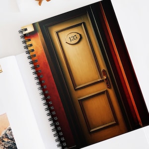 May include: Spiral-bound notebook featuring a stylized image of a wooden door with the number 135. The notebook is open, revealing blank pages and a small photograph. A gold-colored paper clip and sunglasses are also visible.