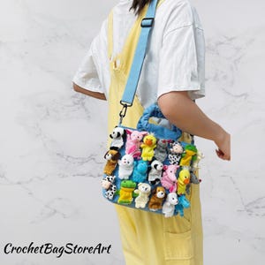 Könnte beinhalten: Eine blaue Tragetasche mit einem hellblauen Schulterriemen, verziert mit zahlreichen kleinen, bunten Fingerpuppen aus Stofftieren. Die Tasche hat einen Griff und der Text "CrochetBagStoreArt" ist unten sichtbar.